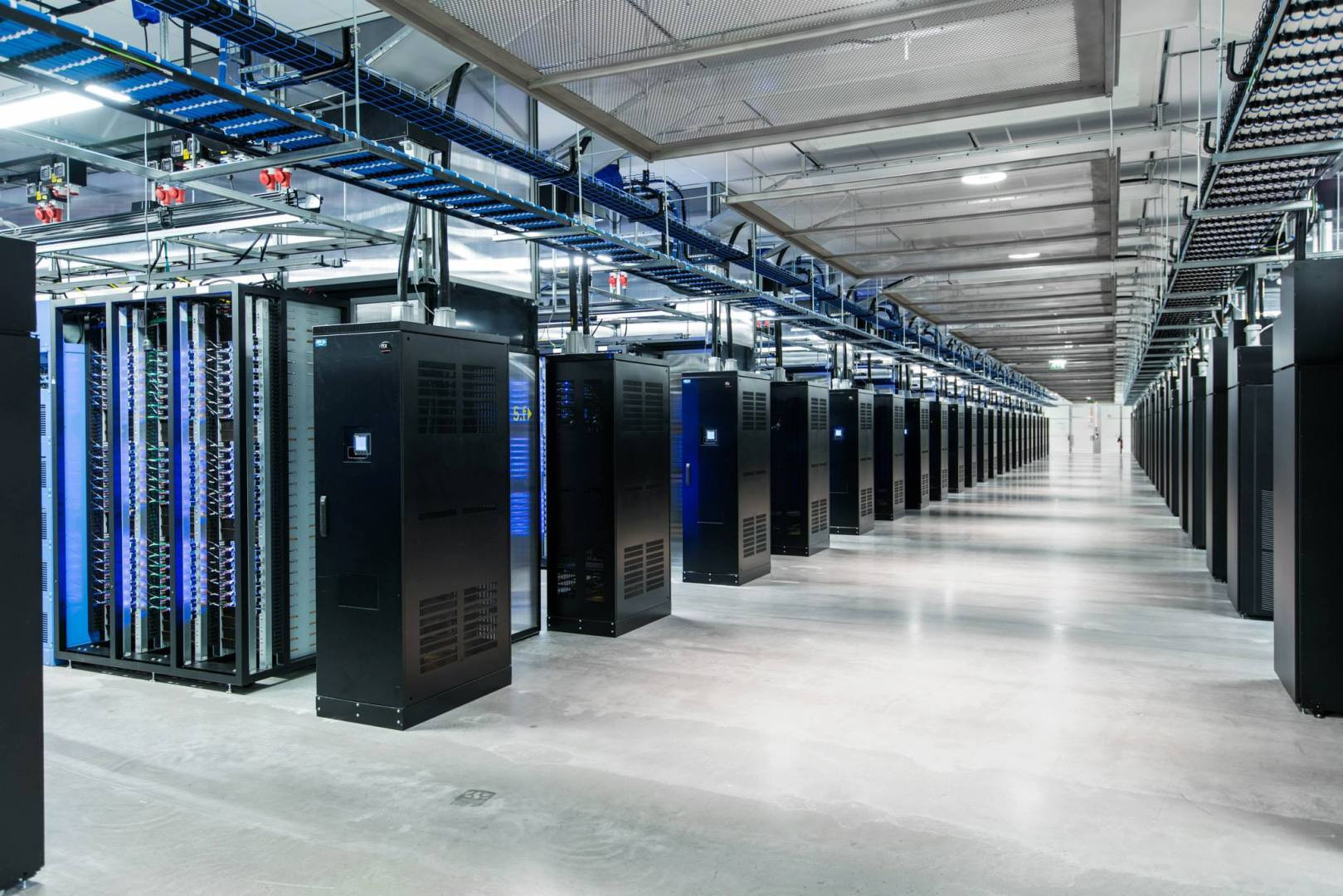  A long room filled with rows of tall black server racks with blinking blue and green lights.